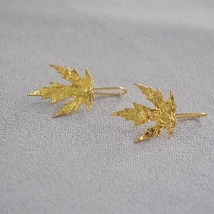 Golden maple leaf earrings with frosted texture on a natural grey fabric background