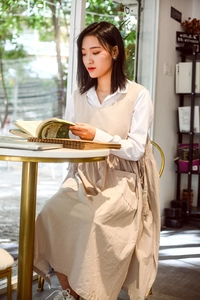 Detail of a beige cotton apron dress worn by a model in a cozy cafe setting