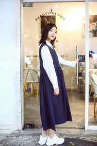 Detail view of the long navy blue sleeveless cotton apron dress over a white long-sleeve shirt