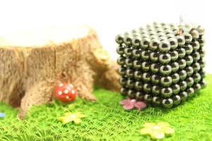 Silver magnetic balls arranged in a cube next to small forest miniatures on green grass