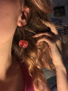 Close-up of red cherry earring with gold hook worn by a model in a natural setting