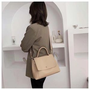 Beige leather handbag with round buckle shown on shoulder of a model in a minimal setting