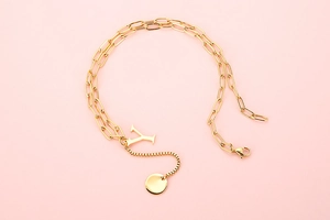 Detail of a gold layered necklace with letter Y pendant and circular tag on pink background