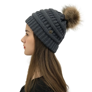 Detail of dark grey cable knit beanie featuring a brown faux fur pom-pom and flower metal tag