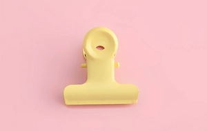 Detailed top view of a pastel yellow metal binder clip on a soft pink background