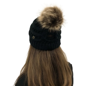 Detail of black knit beanie with large faux fur pompom and small gold logo accent