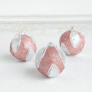 Three pink glitter Christmas ornaments with silver leaves on white textured surface