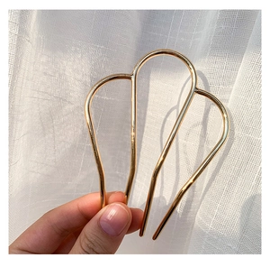 Close-up of a minimalist gold M-shaped metal hair pin held against a soft curtain