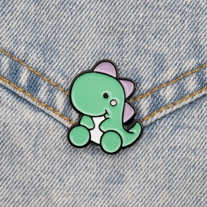 Detail of cute green dinosaur enamel pin attached to light blue denim fabric