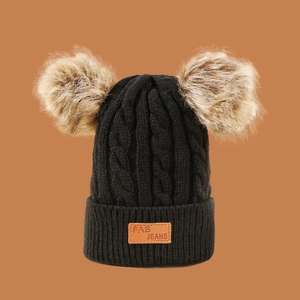 Close-up of black cable knit beanie with dual brown pom-poms and leather logo tag
