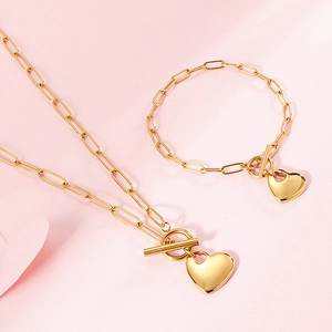 Golden paperclip chain jewelry set with heart pendant and T-bar toggle clasp on pink background