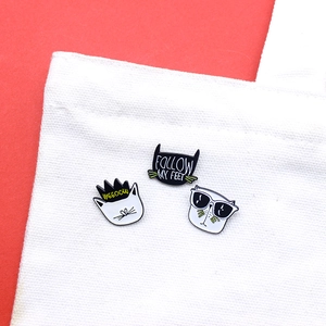 Three cartoon black and white cat enamel pins displayed on a white canvas bag with red background