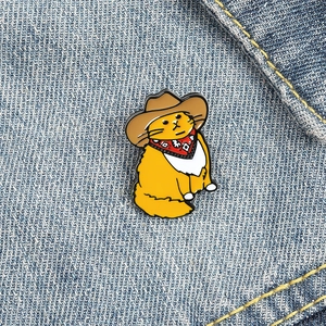 Cartoon orange cat enamel pin wearing a brown cowboy hat and red scarf on denim