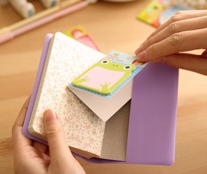 Detail showing hands peeling a green frog sticky note from a purple floral notebook cover