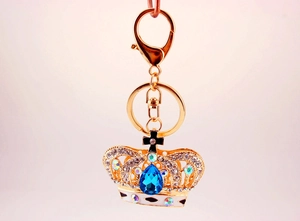 Detail of a golden crown keychain with blue gemstone and crystals on pink background