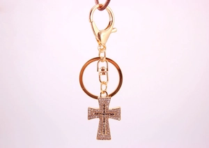 Detail of a golden cross keychain featuring sparkling rhinestones on a soft pink backdrop