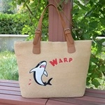 Detail of hand-woven straw tote bag with brown leather straps and shark graphic