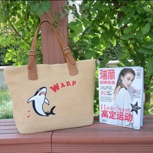 Close-up of beige woven straw tote bag with cartoon shark print and brown leather strap