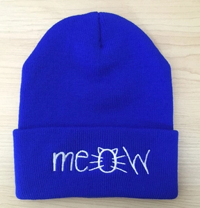 Detail of royal blue knitted beanie featuring white meow embroidery on a flat wood surface