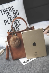 Brown matte crossbody bag with fur pompom charm shown next to a gold iPad for size scale