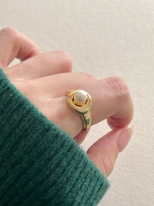 Close-up of a wide gold-tone ring featuring a round white pearl on a textured fabric background
