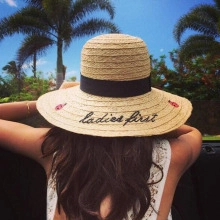 Detail of khaki straw sun hat featuring black ribbon and 'ladies first' embroidery