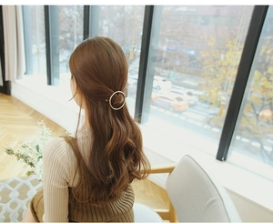 Golden round metal hair clip worn by a woman in a half-up hairstyle by a window