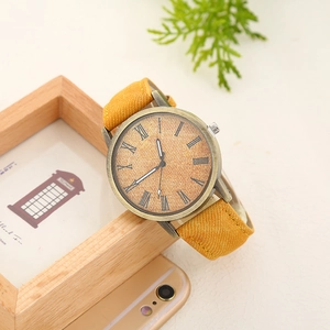 Bronze quartz watch with yellow denim strap and textured dial on a light wood frame