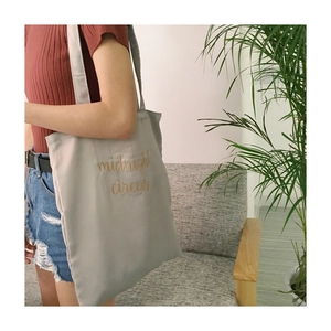 Detail of grey canvas tote bag featuring golden midnight circus text embroidery