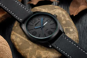 Detalle de reloj militar negro mate con agujas azules y correa de cuero sobre guijarros