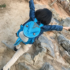 Blue cartoon dinosaur backpack for kids featuring 3D spikes and tail on a rock terrain