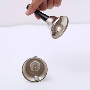 Silver Christmas hand bell with black handle shown in hand and on a flat surface