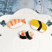 Detailed close-up of three enamel sushi pins on white ceramic plate with shrimp and tamago
