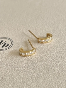 Close-up of gold C-shaped earrings with clear zircon stones on a beige background
