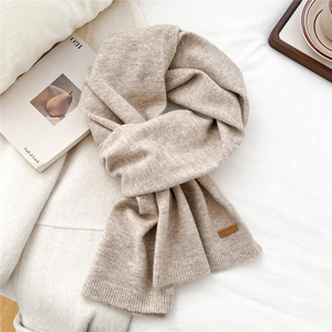 Detail view of the beige knitted scarf featuring a brown leather logo patch and ribbed edge