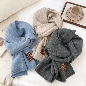Three long solid knit scarves in blue, beige, and grey on a cozy white bed setting