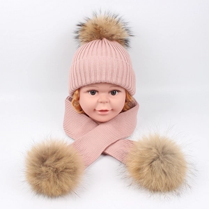 Detail of pink knitted baby hat and scarf set with natural fur pom-poms on grey background