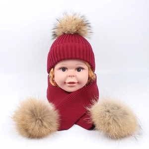 Close-up of red knit baby hat and scarf set with fluffy faux fur pom-poms on white background
