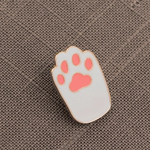 Close-up of a white and pink cat paw enamel pin with gold metal edges on gray fabric