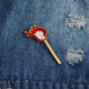 Detailed close-up of a matchstick enamel pin with red flame on blue denim fabric