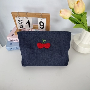 Détail d'une trousse de maquillage en jean avec broderie cerise rouge sur surface blanche