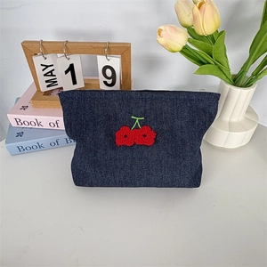 Détail d'une trousse à maquillage en jean bleu avec broderie cerise sur un bureau blanc