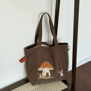 Brown canvas tote bag with mushroom house and bunny embroidery on a wooden stand