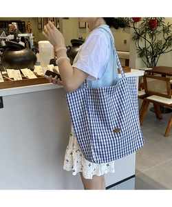 Blue and white checkered canvas tote bag worn by a model in a minimalist cafe setting