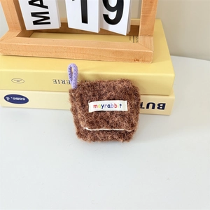 Close-up of brown plush AirPods case featuring a colorful mayrabbit woven label and strap
