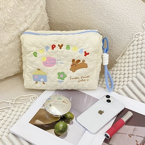 Close-up of cream heart-quilted pouch with puppy embroidery beside makeup on white sofa
