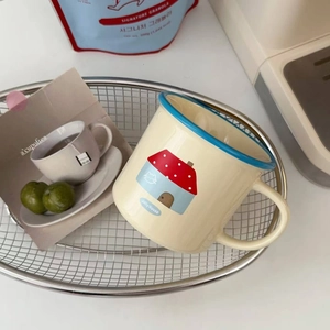 Cream ceramic cartoon mug with blue rim featuring a cat house illustration in a cozy home setting
