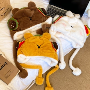 Détail du bonnet peluche ours avec décor burger et liens à pompons sur un lit blanc