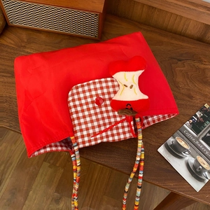 Red plaid tote bag with plush apple charm and beaded handle on a wooden desk