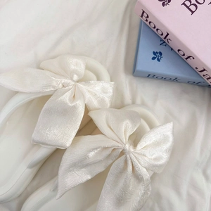 Close-up of white flip-flops with large silk bow details on a soft white fabric surface
